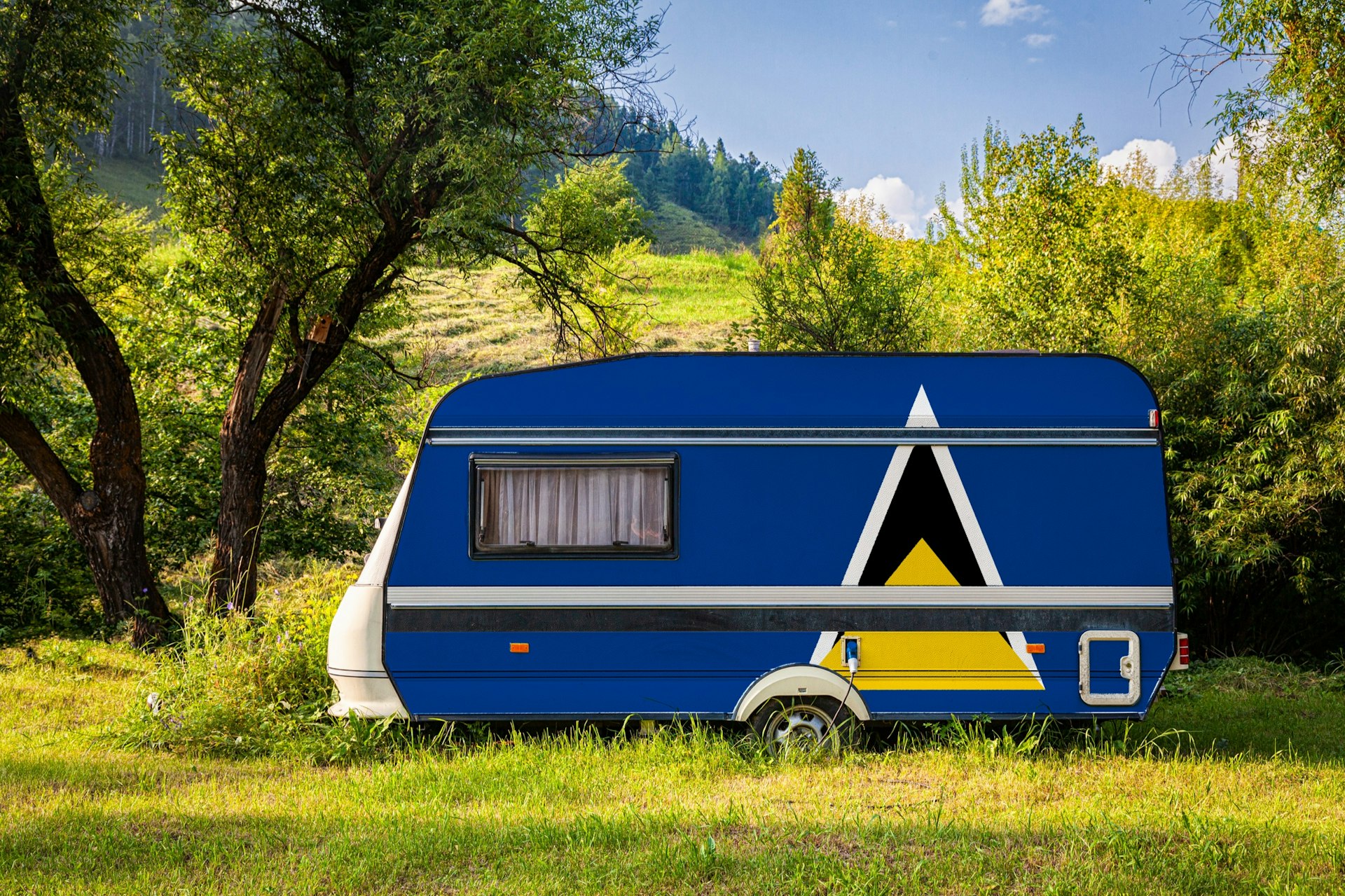 A car trailer, a motor home, painted in the national flag of  Saint Lucia is parked on grass in a mountainous region